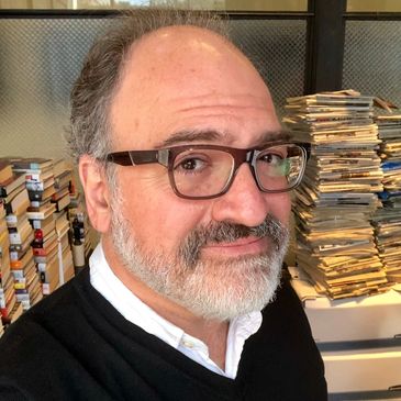 Middle-aged man with glasses and beard in a room filled with stacked books and papers.