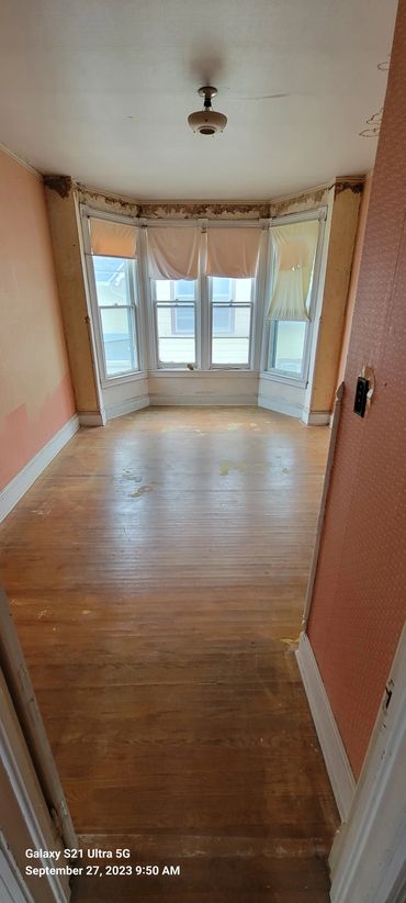 Empty room with damaged walls, worn wooden floor, and partially covered windows.