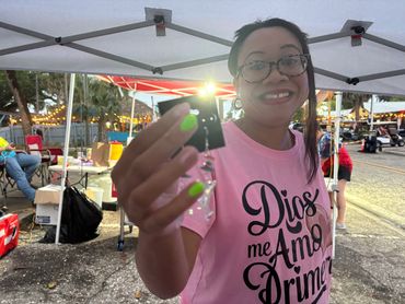 Smiling woman in glasses shows an item at an outdoor event under a canopy.