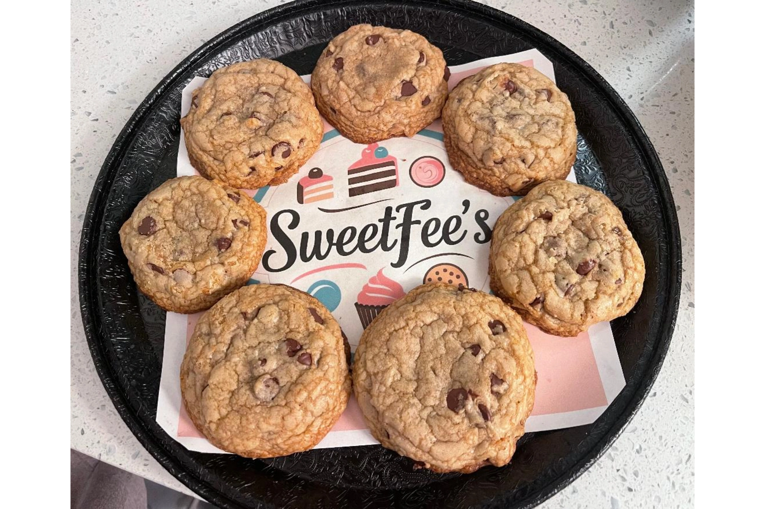 Seven chocolate chip cookies on a SweetFee's branded paper on a black tray.