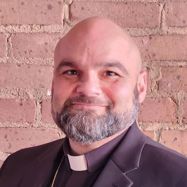 Smiling man with a beard and clerical collar against a brick wall.