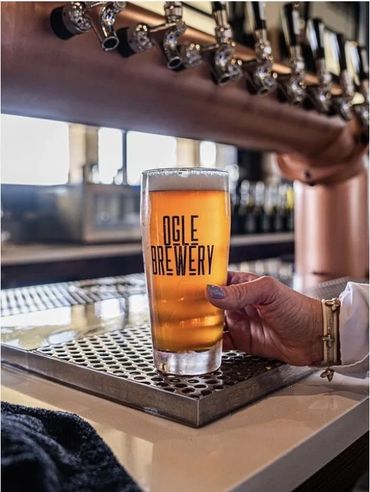 A hand holding a glass of beer from Ogle Brewery at a bar.