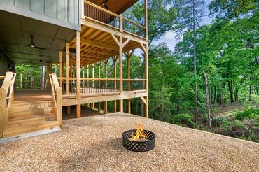Double decker covered back porch with a small staircase & a rocked fire pit area surrounded by trees