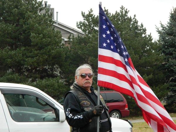 Man in leather vest holding American flag beside a white truck.