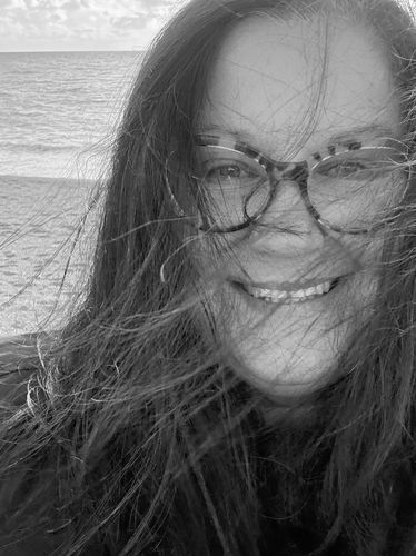 Smiling woman with glasses and windblown hair at the beach during sunset.