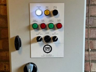 Electrical control panel with indicator lights and switches mounted on a brick wall.