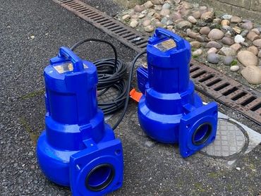 Two blue submersible pumps placed on asphalt near a drain and rocks.