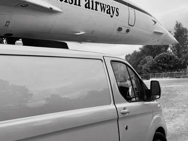 Close-up of British Airways airplane nose over a parked van in grayscale.