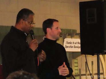 Two men speaking at an event with a microphone and a thank you sign.