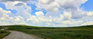 image of gravel backroad with green grass on each side an vast cloud filled sky above