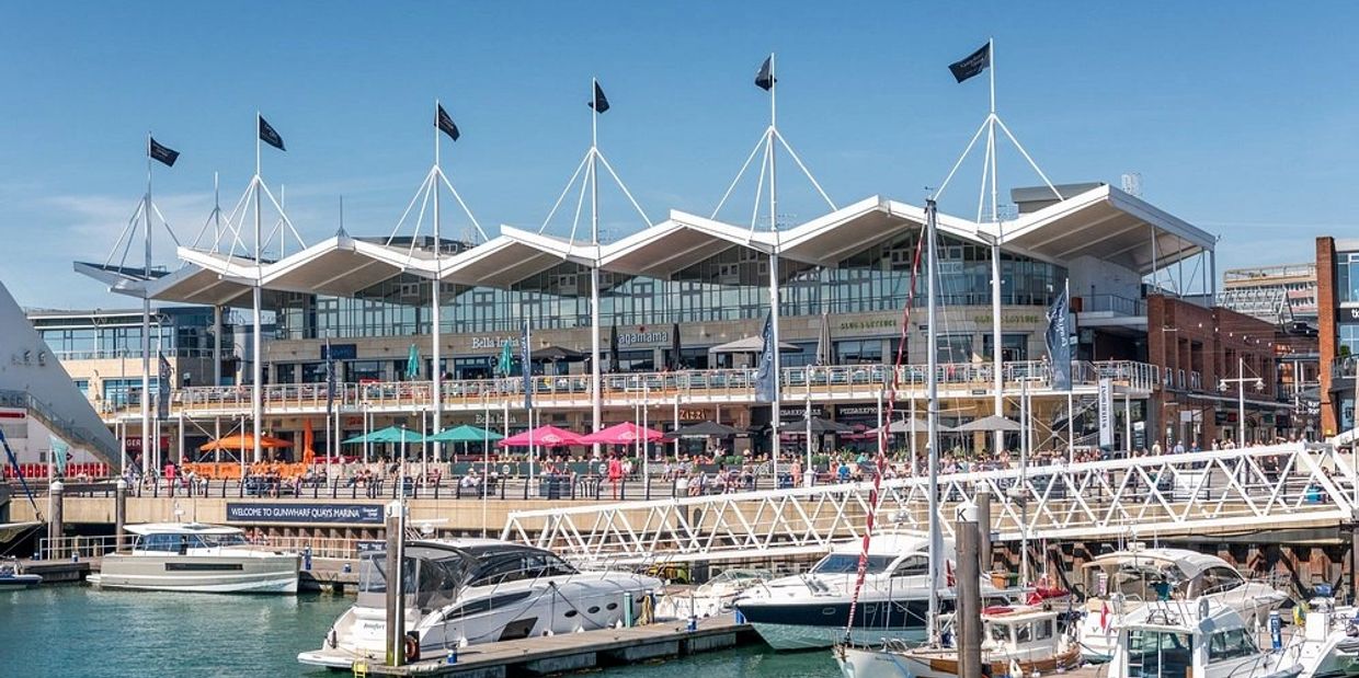 Boats docked at a lively marina with a modern waterfront building and outdoor seating under blue skies.
