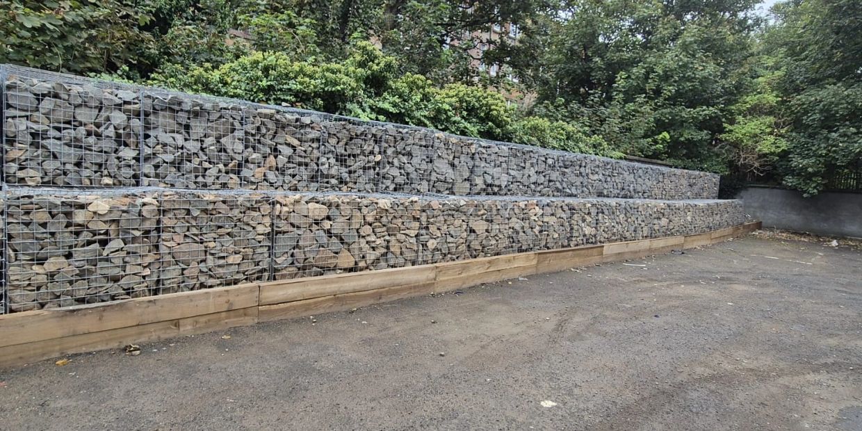Two-tiered rock-filled gabion wall with wooden base and trees in background.