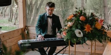 Man in tuxedo playing a Yamaha keyboard in a wooden room with flowers.