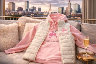 Pink hoodie and white vest with "Bossy Goddess" text displayed on a couch.