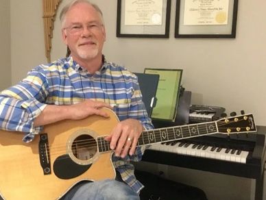 Man who is a chaplain and Certified Music Practitioner sitting with the guitar he uses to provide Live Therapeutic Music to suffering patients