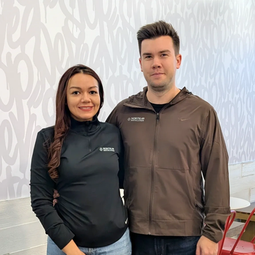 Two people in casual wear posing indoors with a white patterned wall background.