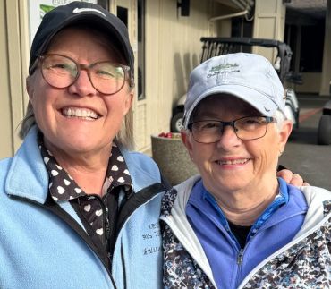 Two smiling elderly women wearing glasses and caps, posing outdoors.