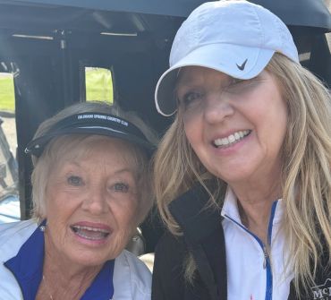 Two women smiling outdoors, one wearing a white cap and the other a visor.
