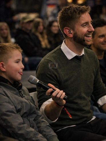Een jongen steekt zijn tong uit terwijl een man lacht en een microfoon vasthoudt.