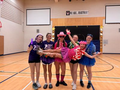 Five girls posing in a gym, one lifted by others in colorful outfits.