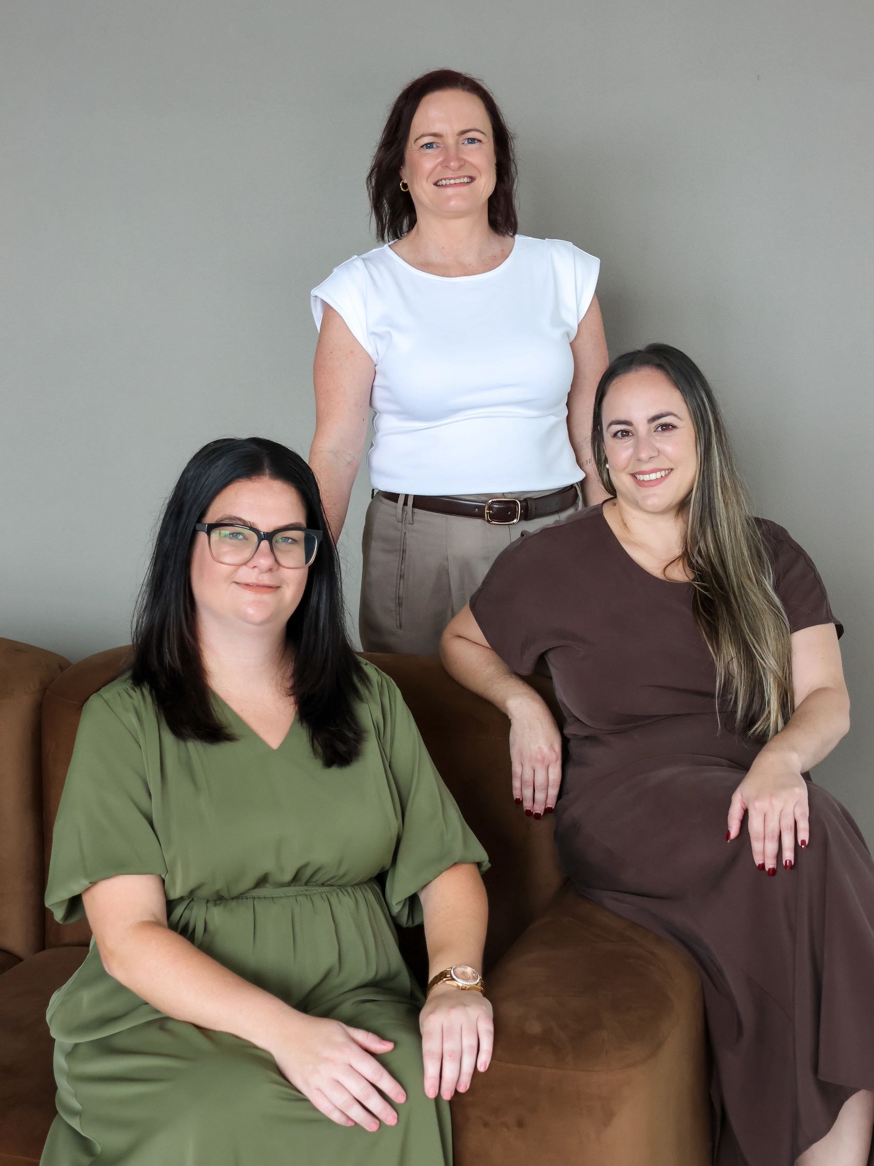 Three smiling women posing indoors against a plain wall.
