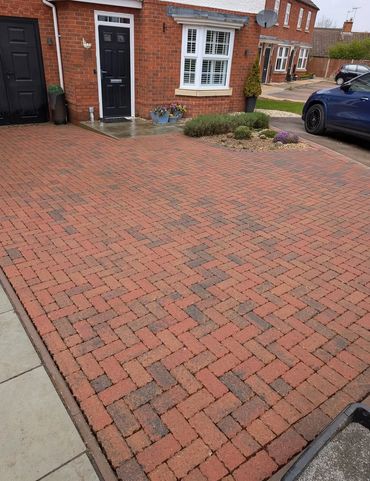 A neat brick driveway in front of a red brick house with black doors and a blue car parked nearby.