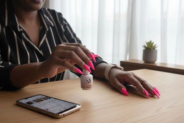 Woman with bright pink nails holding a nail polish bottle at a wooden table.