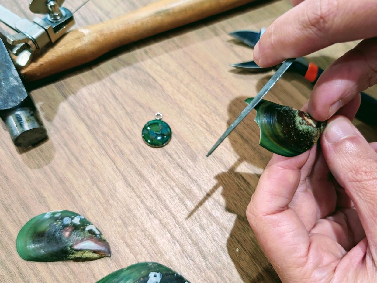 Hands crafting jewelry from green shells with tools on a wooden surface.
