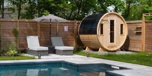 Two lounge chairs under umbrella by swimming pool with wooden barrel sauna.