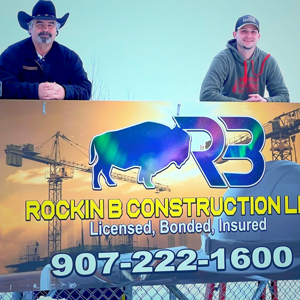 Two men stand behind a construction company sign outdoors in winter.