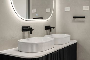 Modern bathroom with dual white sinks and black faucets under a lighted mirror.