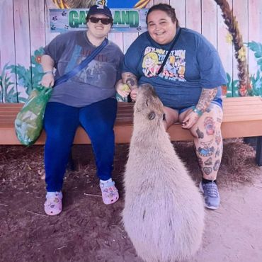 Two people sitting on a bench feeding a capybara under a colorful sign.