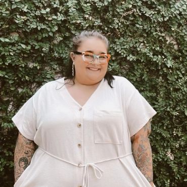 Smiling woman with glasses and tattoos stands against leafy green backdrop.