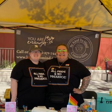 Two smiling individuals wearing matching pride-themed shirts under a yellow tent.