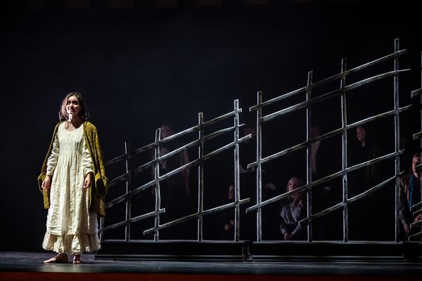 A woman in a long dress stands on stage near a wooden fence with an audience in the background.