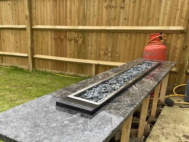 A long granite countertop with a rectangular fire pit insert, supported by wooden legs in a backyard.