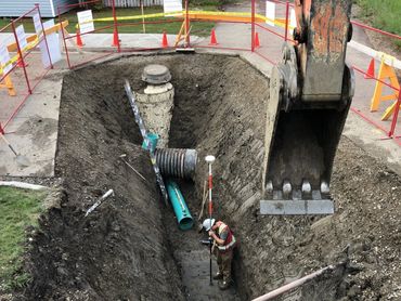 Worker inside a deep excavation site with heavy machinery and pipes.