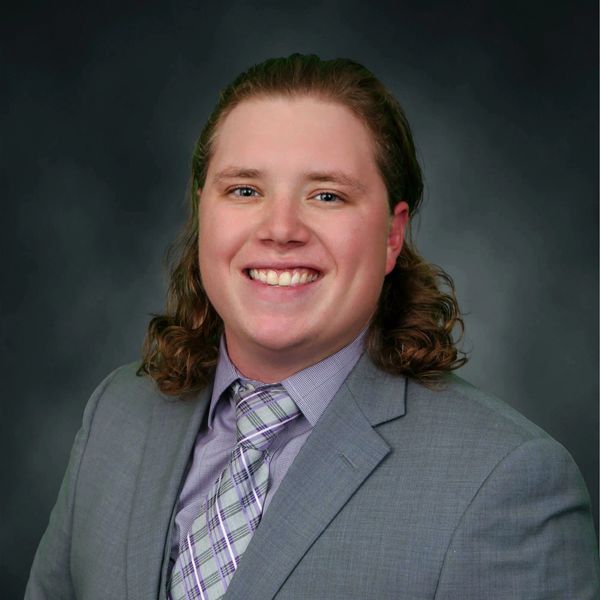 A smiling man in a gray suit and plaid tie against a dark background.