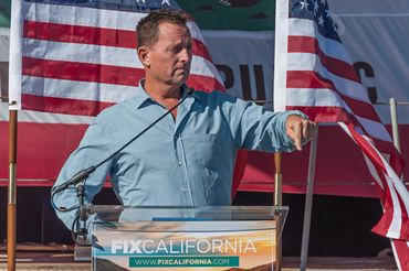 Ambassador Ric Grenell, giving speech in front of fixcalifornia.com sign and audience.