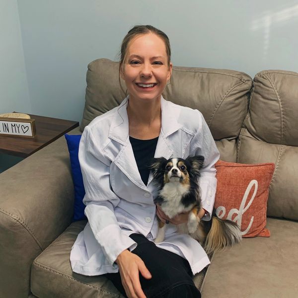 Smiling woman in a white coat holding a small dog on a couch.