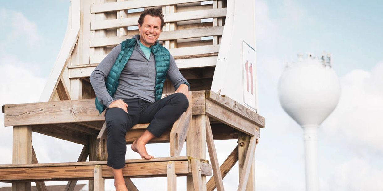 A man in casual clothes sitting barefoot on a wooden lifeguard tower under a clear sky.