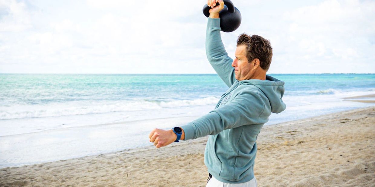 Man lifting a kettlebell on a sandy beach near the ocean.