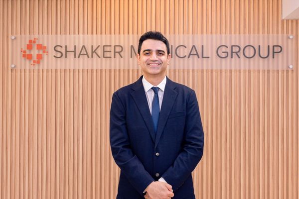 Man wearing a blue suit stands in front of the sign "Shaker Medical Group" behind him on the wall.