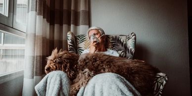 Senior lady with a oxygen mask on while she sits on the couch with her dog on her lap.