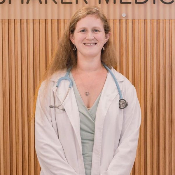 Julie Hedlund, FNP -C, RN, wearing a white coat stands in front of a Shaker Medical Group sign.
