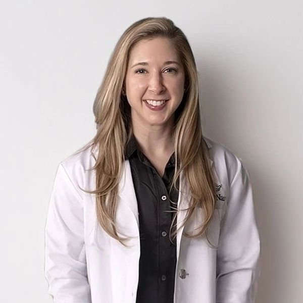 RN Christina Masklee wearing a white coat stands against a white wall.