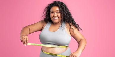 Smiling black overweight woman in sportswear checking waist with measuring tape, isolated on pink.