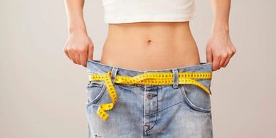 A close up of a woman's flat stomach with loose pants and a measuring tape for a belt.