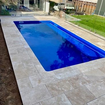 A modern rectangular blue pool with beige stone tiles around it in a backyard.