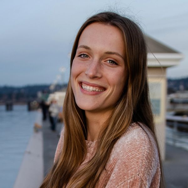 Smiling woman with long hair standing near a waterfront at dusk.